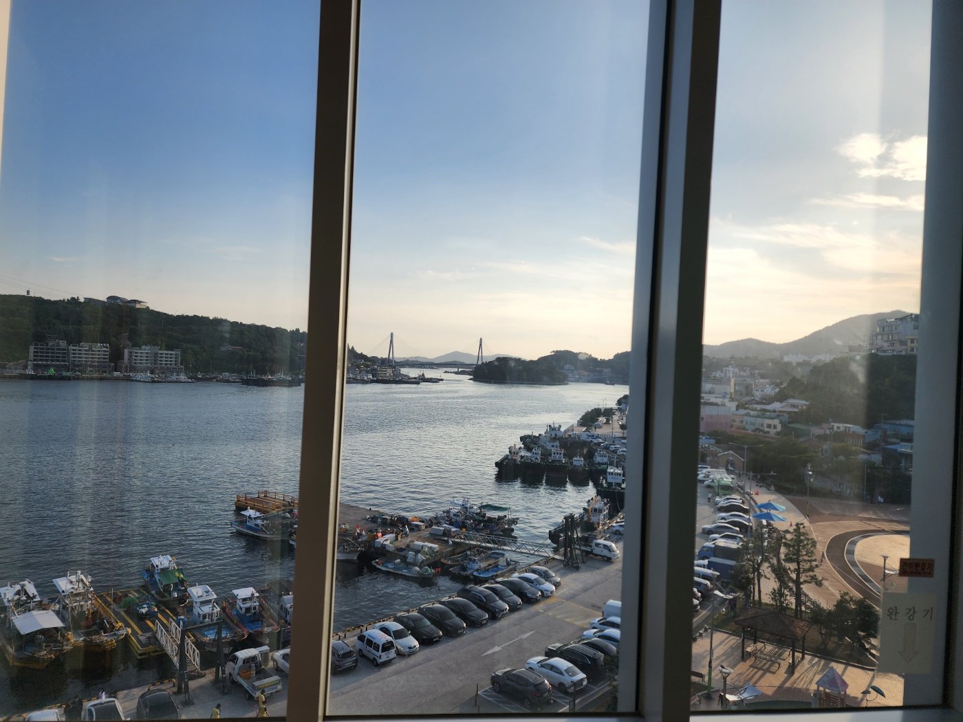건물 창가에서 내려다본 여수 항구 — 멀리 사장교와 정박한 어선, 주차장